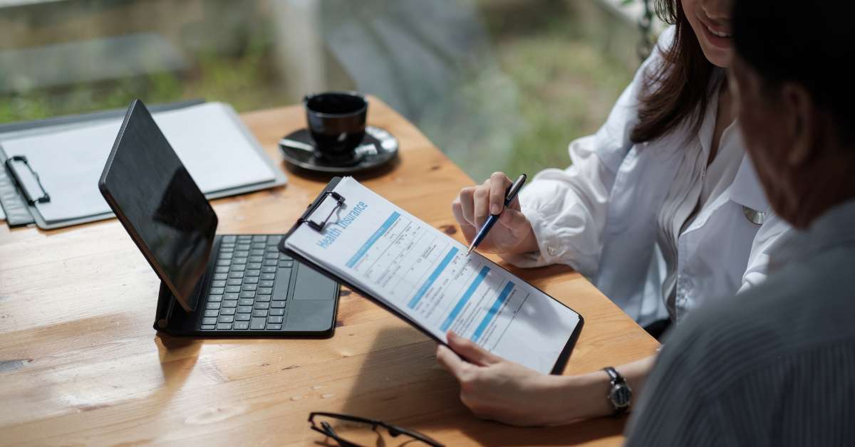 doctor sitting in front of a laptop and consulting a clipboard with another person